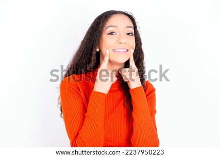Strong healthy straight white teeth. Close up portrait of happy teen girl with curly hair wearing red sweater over white background with beaming smile pointing on perfect clear white teeth.
