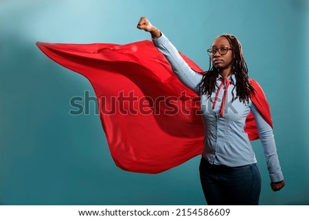 Strong justice defender with superpowers looking at camera with fist in air. Brave and proud looking young adult superhero woman acting like a flying hero while wearing mighty cape on blue background.