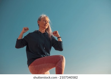 Strong Lady In Pink Leggins And Blue Hoodie Exercising , Lifting One Knee Up And Looking Ahead On Sunny Day