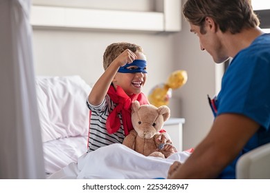 Strong little boy wearing superhero blue mask and red cape sitting on hospital bed playing with nurse. Happy superhero kid gesturing with playful doctor at clinic. Happy child playing as superman. - Powered by Shutterstock
