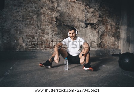 Strong male athlete in sportswear and sneakers looking away and relaxing with water bottle while sitting on floor with headphones and listening music during break of intense workout in gym