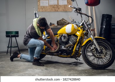 A Strong Male Biker Repairs His Motorcycle In The Garage. Interior Of The Workshop