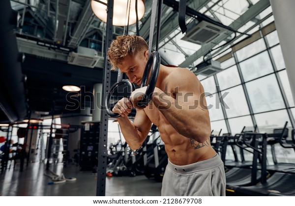 Strong\
man doing exercise on to gymnastic rings at gym. Fit male athlete\
training on gymnastic rings in light sport\
hall