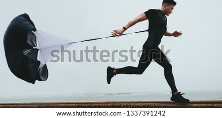 Strong man doing workout using resistance parachute outdoors. Young asian man in black sports clothing running with parachute.