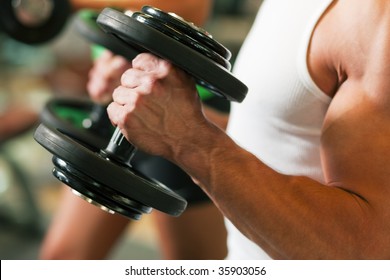 Strong Man Exercising With Dumbbells In A Gym, In The Background A Woman Also Lifting Weights; Focus On Hands