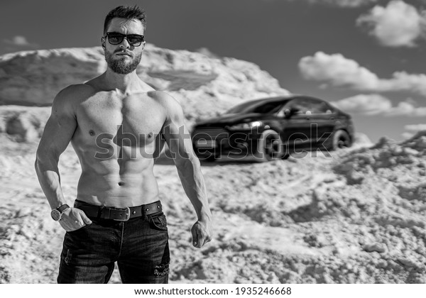 Strong man with muscular body\
type. Monochrome photo of bodybuilder in sunglasses standing in\
front of modern car. Beautiful location with white mountains and\
sky.
