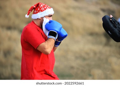 A strong man with a Santa Claus hat and beard, dressed in a red T-shirt and with blue boxing gloves on his hands, stands in a boxer pose and prepares to strike. On open air. - Powered by Shutterstock