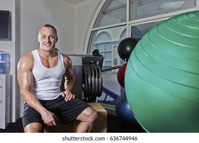 Strong Man With Smilling Face, Inside Room Of A Luxury Gym, Heavy Exercise Tools Near Wall, A Body Builder Sitting On A Bench, Perfect Lights And Designs.