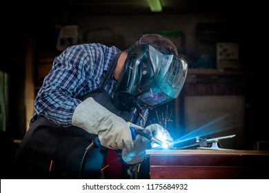 A strong man welder in a black T-shirt, in a welding mask and welders leathers weld metal welding machine in the workshop, in the sides fly the blue sparks