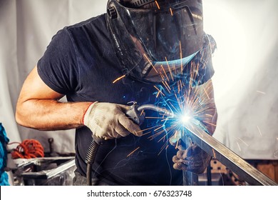 A strong man is a welder in a black T-shirt, in a welding mask and welders leathers, a metal product is welded with a welding machine in the garage, blue sparks fly to the sides - Shutterstock ID 676323748