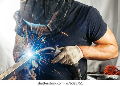 A strong man welder in a black T-shirt, in a welding mask and welders leathers weld metal welding machine in the workshop, in the sides fly the blue sparks
