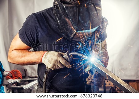 A strong man is a welder in a black T-shirt, in a welding mask and welders leathers, a metal product is welded with a welding machine in the garage, blue sparks fly to the sides