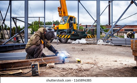 A strong man is a welder in brown uniform, welding mask and welders leathers, a metal product is welded with a arc welding machine at the construction site, blue sparks fly to the sides