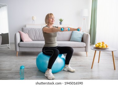 Strong mature woman working out with dumbbells on fitness ball at home, blank space. Fit senior lady taking care of her health, having domestic training, leading active lifesyle, indoors