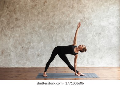 Strong middle-aged woman practicing yoga, standing in Utthita Trikonasana pose, extended triangle exercise, working out at home or in yoga studio. Mindfulness and healthy lifestyle concept, copy space