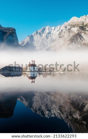 Strong morning fog over church St. Bartholomew on Konigsee lake during winter time. Beautiful rocky alps on background with clear reflection on water.