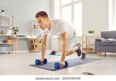 Strong, motivated, determined, young man in sportswear having a fitness workout at home, holding dumbbells, and doing a plank exercise with knee tucks on a rubber sports mat - Powered by Shutterstock