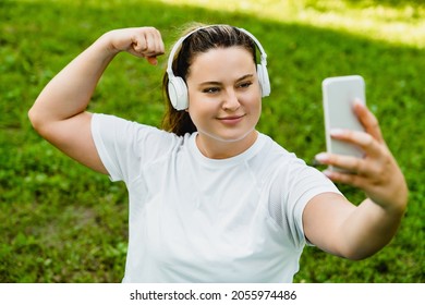 Strong muscular fit plump plus-size caucasian woman athlete taking selfie photo image, having video call conversation with trainer, posting on social media outdoors in park
