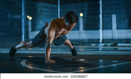 Strong Muscular Fit Shirtless Young Man is Doing One-Hand Push Up Exercises. He is Doing a Workout in a Fenced Outdoor Basketball Court. Night After Rain in a Residential Neighborhood Area. - Powered by Shutterstock