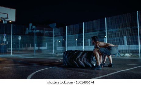 Strong Muscular Fit Young Shirtless Man Is Doing Exercises In A Fenced Outdoor Basketball Court. He's Flipping A Big Heavy Tire In A Foggy Night After Rain In A Residential Neighborhood Area.