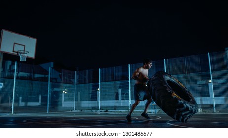 Strong Muscular Fit Young Shirtless Man Is Doing Exercises In A Fenced Outdoor Basketball Court. He's Flipping A Big Heavy Tire In A Foggy Night After Rain In A Residential Neighborhood Area.