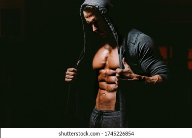 Strong muscular fitness model body six pack abs posing. Brutal bodybuilder fitness model posing in gym on black background.  Fitness man wearing hoodie on naked body.