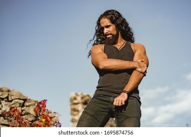 Strong Muscular Handsome Man At Blue Summer Sky Background. Fashionable Man With Long Wavy Hair And Strong Healthy Body. Mens Style And Wellness Concept