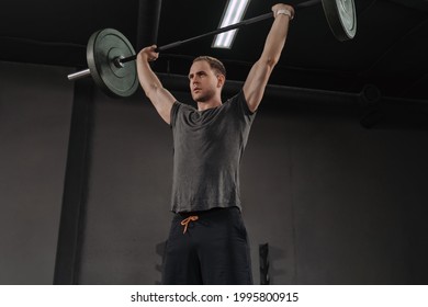 Strong muscular man holding heavy barbell overhead at crossfit gym, doing military press exercises as a part of functional training. Weightlifter exercising with barbell. 