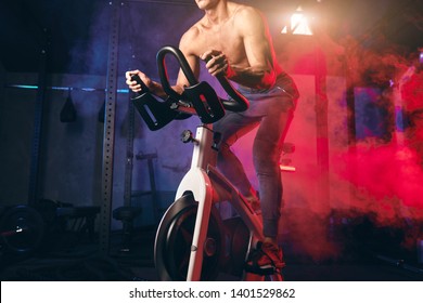 Strong muscular white athletic man shirtless training in a bicycle simulator in the gym, working out enduarance and enhancing his sporting spirit and body.