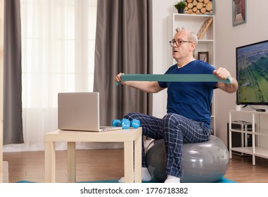 Strong Old Man Training Online With Resistance Band. Old Person Pensioner Online Internet Exercise Training At Home Sport Activity With Dumbbell, Resistance Band, Swiss Ball At Elderly Retirement Age.