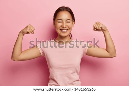 Strong powerful Asian woman with dark combed hair, toothy smile, raises arms and shows biceps, has piercing in ear, wears casual rosy t shirt, models against pink background. Look at my muscles!