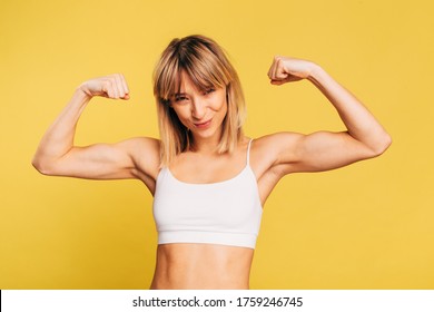 Strong Powerful And Slim Young Woman Hold Hands Up And Posing On Camera. Well-built Fit Model Showing Her Strenght In Muscles. Isolated Over Yellow Background