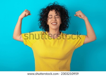 Strong powerful young arab woman wearing yellow T-shirt over blue background toothy smile, raises arms and shows biceps. Look at my muscles!