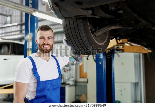 Strong repairman cheerful young male professional\
technician mechanic man wears denim blue overalls white t-shirt\
stand near car lift changes wheel tires work in vehicle repair shop\
workshop indoors