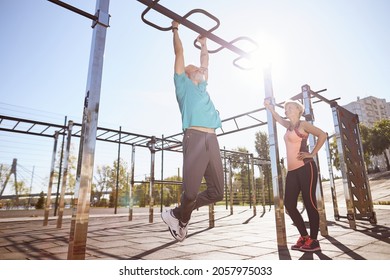 Strong senior man in sportswear doing pull-ups on horizontal bar while training with his wife outdoors. Full length. Beautiful aged couple exercising in the morning - Powered by Shutterstock