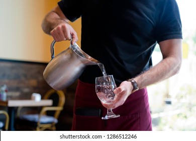 Strong Slim Man With Muscular Muscles Working As A Waiter In A Restaurant In A Black T-shirt And Burgundy Apron Pours Water From A Stainless Steel Jug Into A Customers Glass