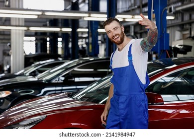 Strong Smiling Friendly Happy Young Male Professional Technician Car Mechanic Man Wears Denim Blue Overalls White T-shirt Wave Hand Calling Work In Light Modern Vehicle Repair Shop Workshop Indoors