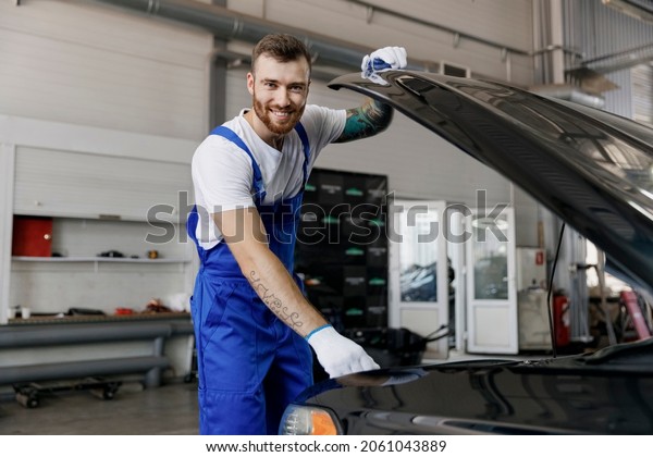 Strong smiling happy troubleshooter young male\
professional technician car mechanic man in denim blue overalls\
white t-shirt raised hood bonnet work in light modern vehicle\
repair shop workshop\
indoor