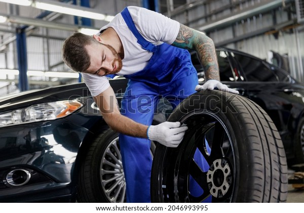 Strong smiling happy young male professional\
technician car mechanic man 20s wears denim blue overalls white\
t-shirt stand hold check wheel work in light modern vehicle repair\
shop workshop indoors