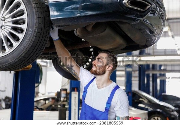 Strong smiling happy young male professional\
technician mechanic man wears denim blue overalls white t-shirt\
stand near car lift check technical condition work in vehicle\
repair shop workshop\
indoors