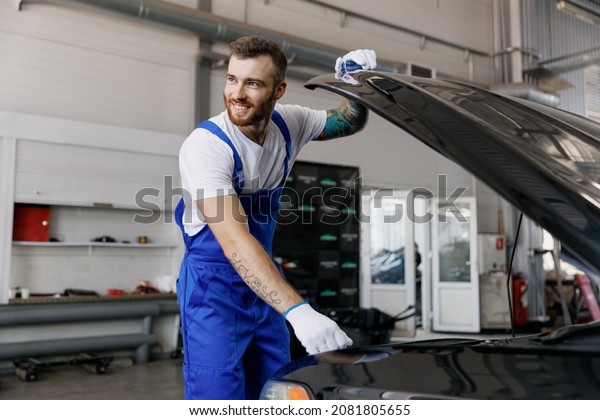 Strong smiling troubleshooter young male\
professional technician car mechanic man in denim blue overalls\
white t-shirt raised hood look aside work in light modern vehicle\
repair shop workshop\
indoor