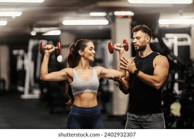 A strong sportswoman is doing exercises with dumbbells in a gym while her instructor helping her. Gym workout. Fitness trainer. - Powered by Shutterstock