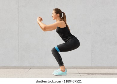 Strong sporty Caucasian woman has exercises with rubber resistance band, trains legs, works on muscles, dressed in t shirt and leggings, stands indoor against grey background in fitness studio