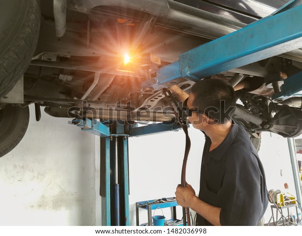 Strong technical welder short hair man wearing protect\
black glasses using a fire gas welding exhaust steel tube under\
hoist lifting car. Background for safety first in working or\
specialist worker. 