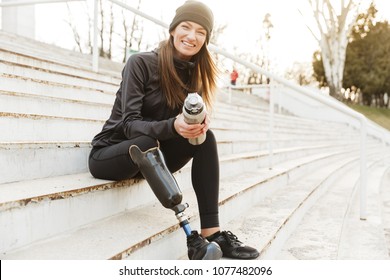 Strong willed handicapped woman in black tracksuit with prosthetic leg laughing while sitting at the street stairs with thermos cup in hands - Powered by Shutterstock