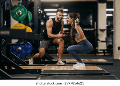 A strong woman doing squats with bar in a gym while her trainer is supporting her. - Powered by Shutterstock