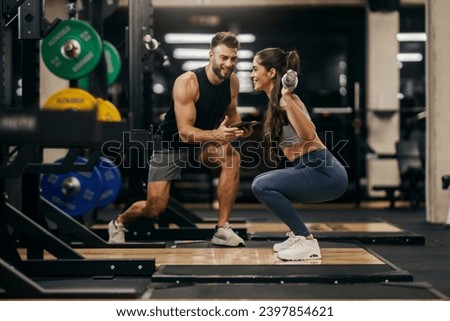 A strong woman doing squats with bar in a gym while her trainer is supporting her.