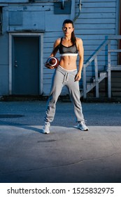 Strong Woman Holding A Football. Urban Street Setting. She Is Wearing A Tank Top And Sweat Pants.