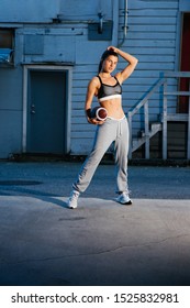 Strong Woman Holding A Football. Urban Street Setting. She Is Wearing A Tank Top And Sweat Pants.