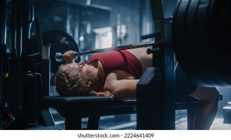 A Strong Woman Lifting Extremely Heavy Weights on a Barbell and Working Out. A Resolute Female Bodybuilder Training Alone in a Hardcore Dark Gym, Doing Bench Press Exercises - Powered by Shutterstock